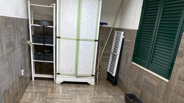 A room with a herringbone-patterned floor, a metal chair, a white cabinet with shelves, and a window with green shutters. - The Chiaia steps :camera white (Napoli)