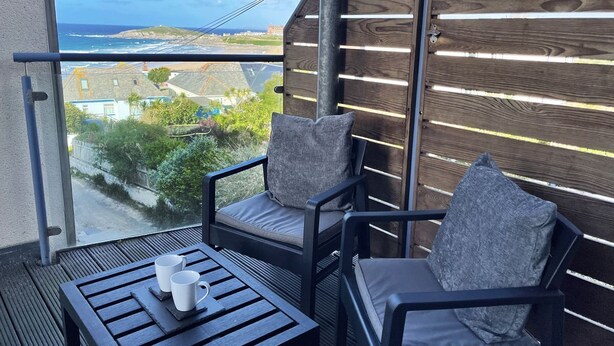 Ein Balkon mit zwei Stühlen und einem Tisch, der Blick auf die Küste mit Gebäuden und viel Grün bietet.