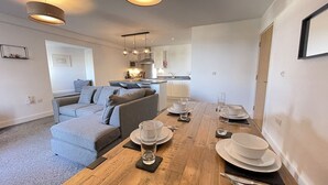 Ein modernes Wohnzimmer mit einem Holzespeisetisch, der für vier Personen gedeckt ist, einem grauen Sofa und einer Küche im Hintergrund.