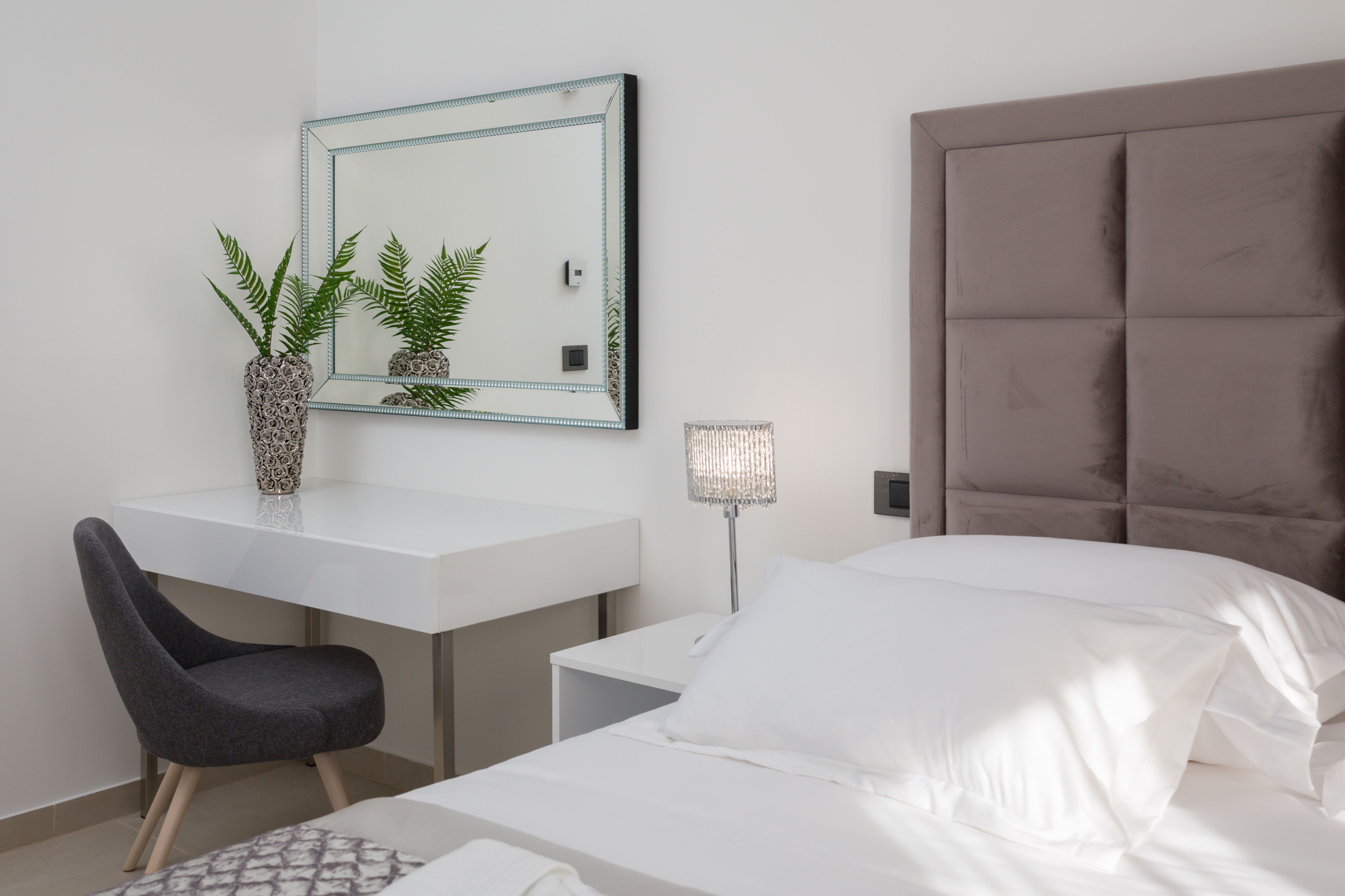 Una habitación de hotel moderna con una cama blanca, un escritorio con una silla, un espejo grande y una mesita de noche con una lámpara.