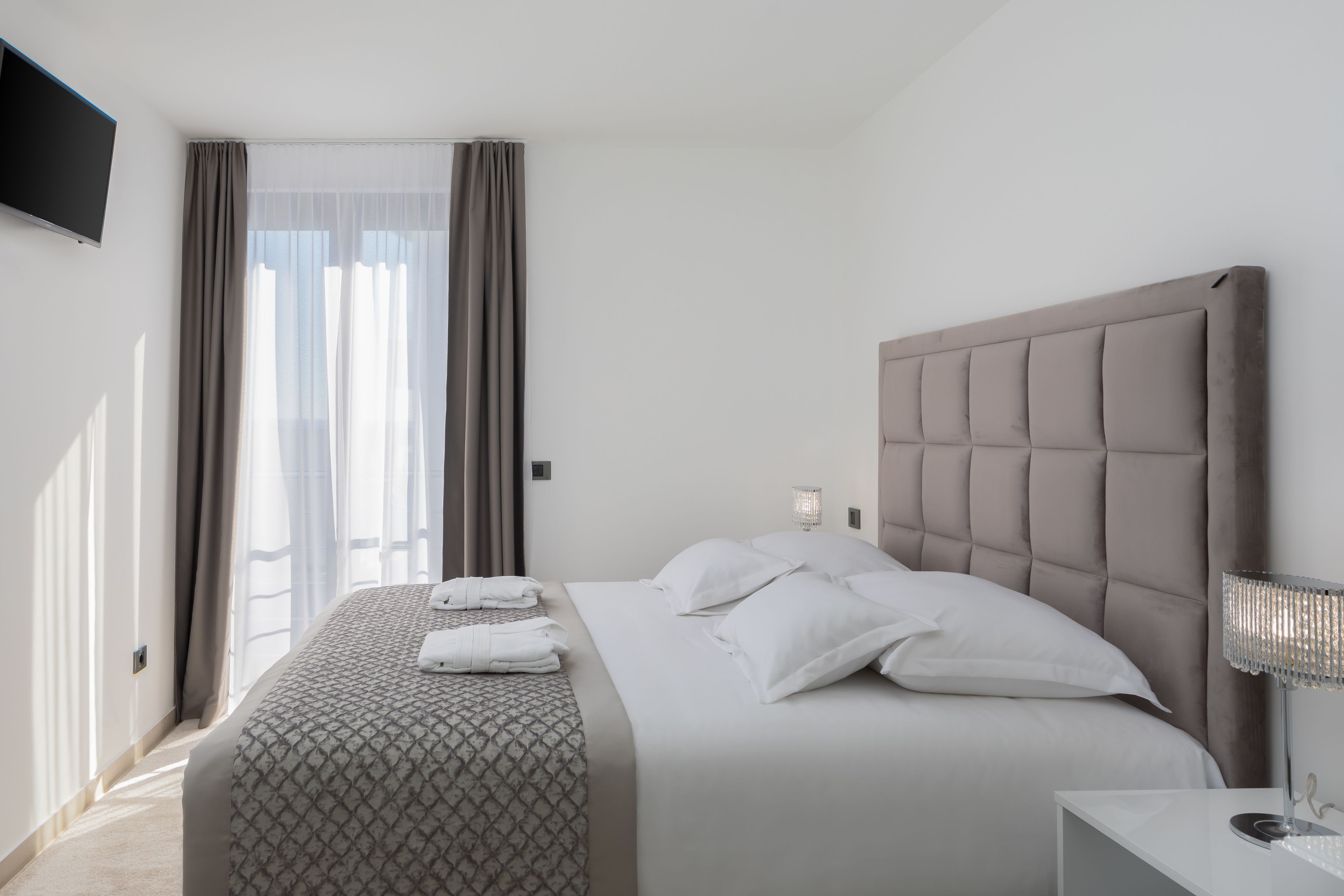 A hotel room with a large bed, a flat-screen TV mounted on the wall, and a window with curtains.