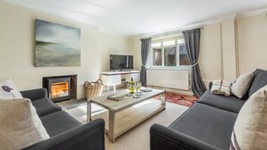 A modern living room with a fireplace, a flat-screen TV, and a large window with curtains. - The Tack House, Holkham, Norfolk (Wells-Next-The-Sea)