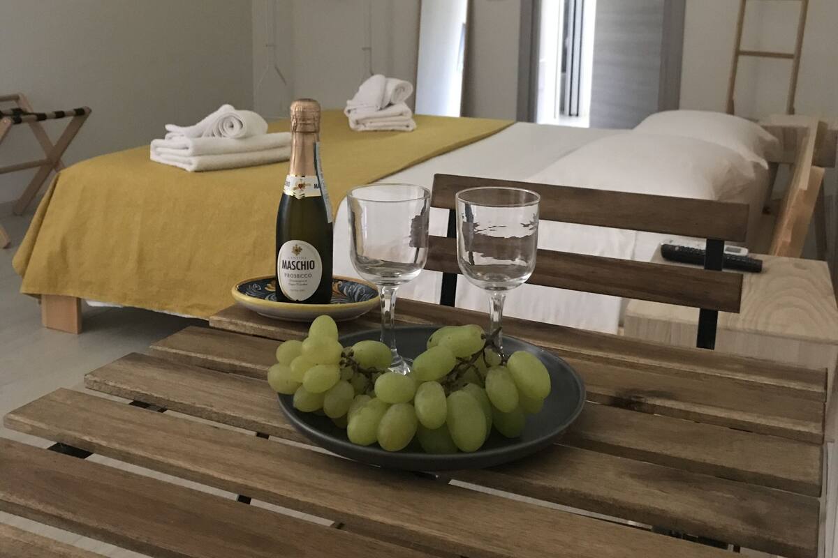Une chambre d’hôtel avec un lit, une table de chevet sur laquelle se trouve une bouteille de Maschio Prosecco, deux verres et une assiette de raisins verts.