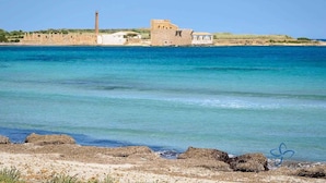 A coastal scene with a historic building complex by the sea, featuring stone walls and a chimney. - Le Camere di Don Vito (Noto)