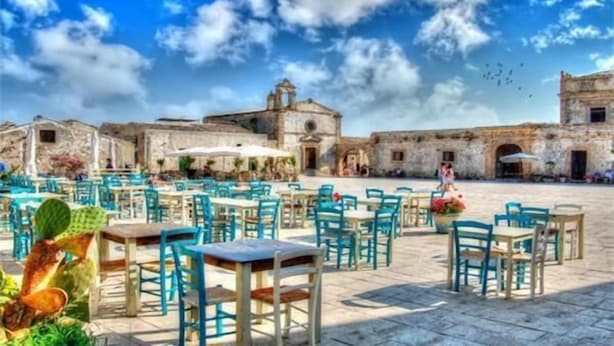 An outdoor café with tables and chairs, a historic building in the background, and a clear blue sky. - Le Camere di Don Vito (Noto)