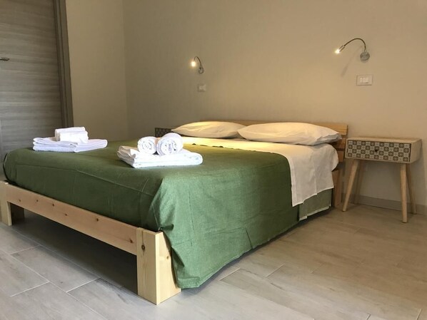 Una cama individual con una colcha verde, almohadas blancas y toallas dobladas cuidadosamente a un lado. Hay dos lámparas de pared a cada lado de la cama.