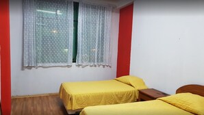 Une chambre avec deux lits, chacun recouvert d’une couvre-lit jaune, une fenêtre ornée de rideaux à motifs et un mur rouge.