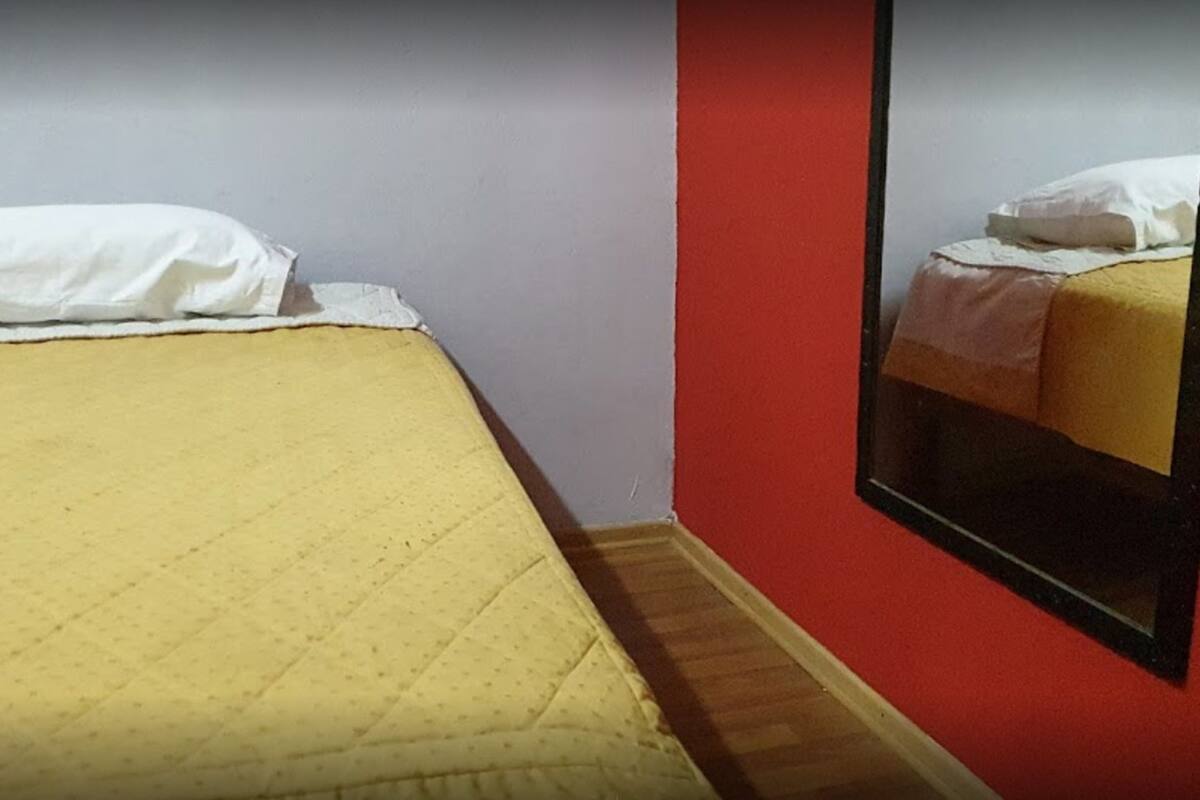 A single bed with a yellow bedspread and white pillows in a room with a red wall and a mirror reflecting the bed.