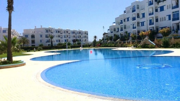 A large, clear swimming pool surrounded by a paved walkway, with white buildings and palm trees in the background. - Jaharasmir Residence 2905 (Mdiq)