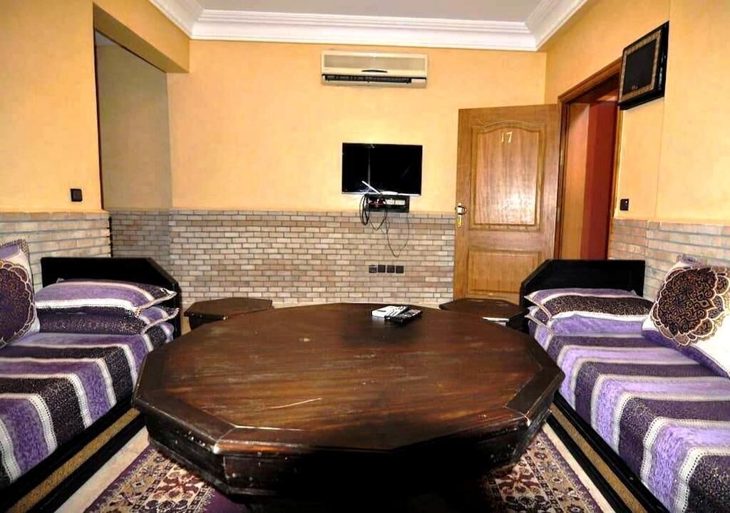 A conference room with a large wooden table, two sofas with patterned cushions, a television mounted on the wall, and a door with a number plate.