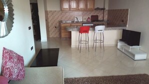 A living room with a sofa, a coffee table, and a TV stand. There is a kitchen area with cabinets, a microwave, and two bar stools. - Atlantic Ocean Residence 3040 (Asilah)