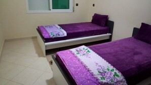 Two single beds with purple bedding in a room with a window and a green screen. - Atlantic Ocean Residence 3040 (Asilah)