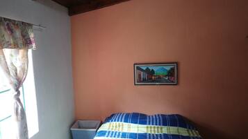 A bedroom with a bed, a trash bin, a window with curtains, and a framed picture on the wall.