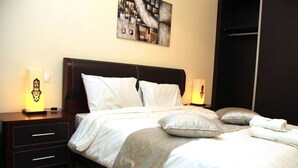 A neatly made bed with white bedding and a grey throw, a dark wooden nightstand with a lamp, and a modern abstract painting on the wall. - The Greens Residence 3621 (SAIDIA)