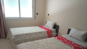 Una habitación con dos camas, cada una con una colcha estampada y un detalle rojo. Hay una ventana con cortinas, y las paredes son lisas.