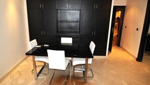 Uma sala de jantar com mesa preta e cadeiras brancas, um armário preto com gavetas, e um corredor que dá para outro quarto.