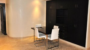 Uma área de refeições moderna com mesa preta e cadeiras brancas, um armário preto com gavetas e piso em tons claros.