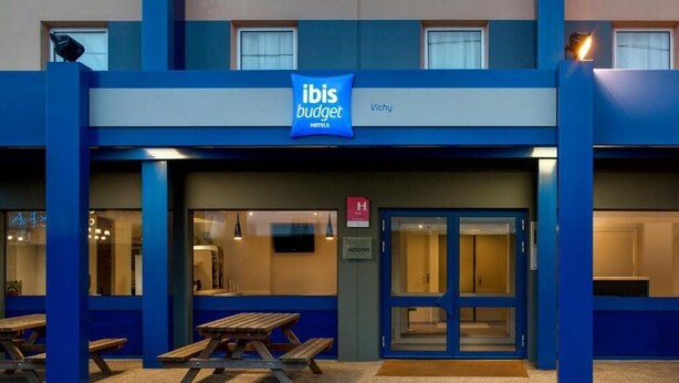 Une entrée d’hôtel moderne aux couleurs bleu et blanc, une enseigne indiquant les hôtels ibis budget, et un comptoir d’accueil doté d’une porte en verre.