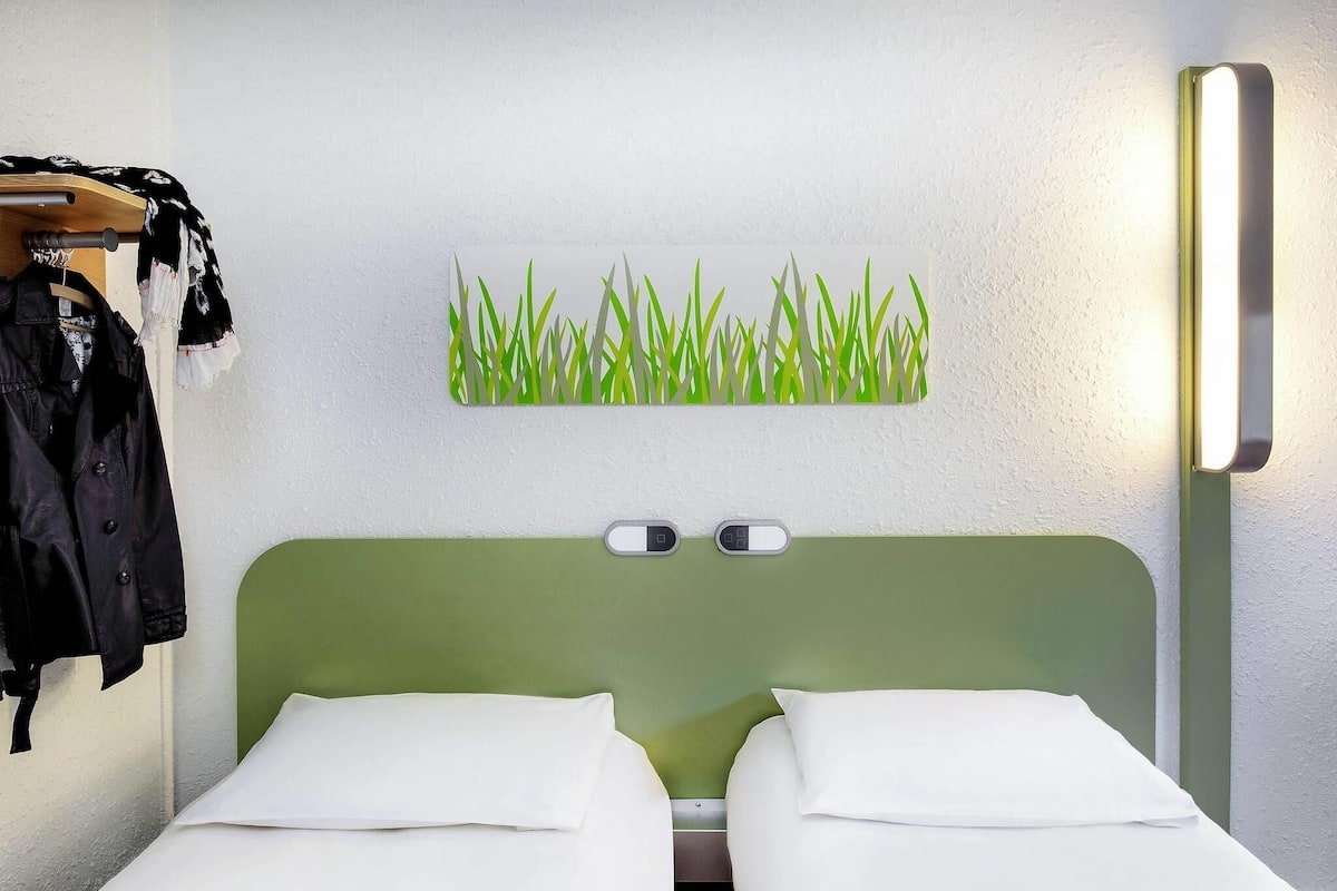 A hotel room with two single beds, a green headboard, a wall-mounted light, and a coat rack with a jacket.