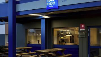 Een hotelgevel met een bord waarop 'ibis budget hotels' en 'Vichy' staat. Er zijn terrassen met houten tafels en banken.