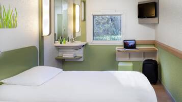 Et hotelværelse med seng, skrivebord med en bærbar computer, tv, et vindue med udsigt til træer og et badeværelse med spejl og vask.