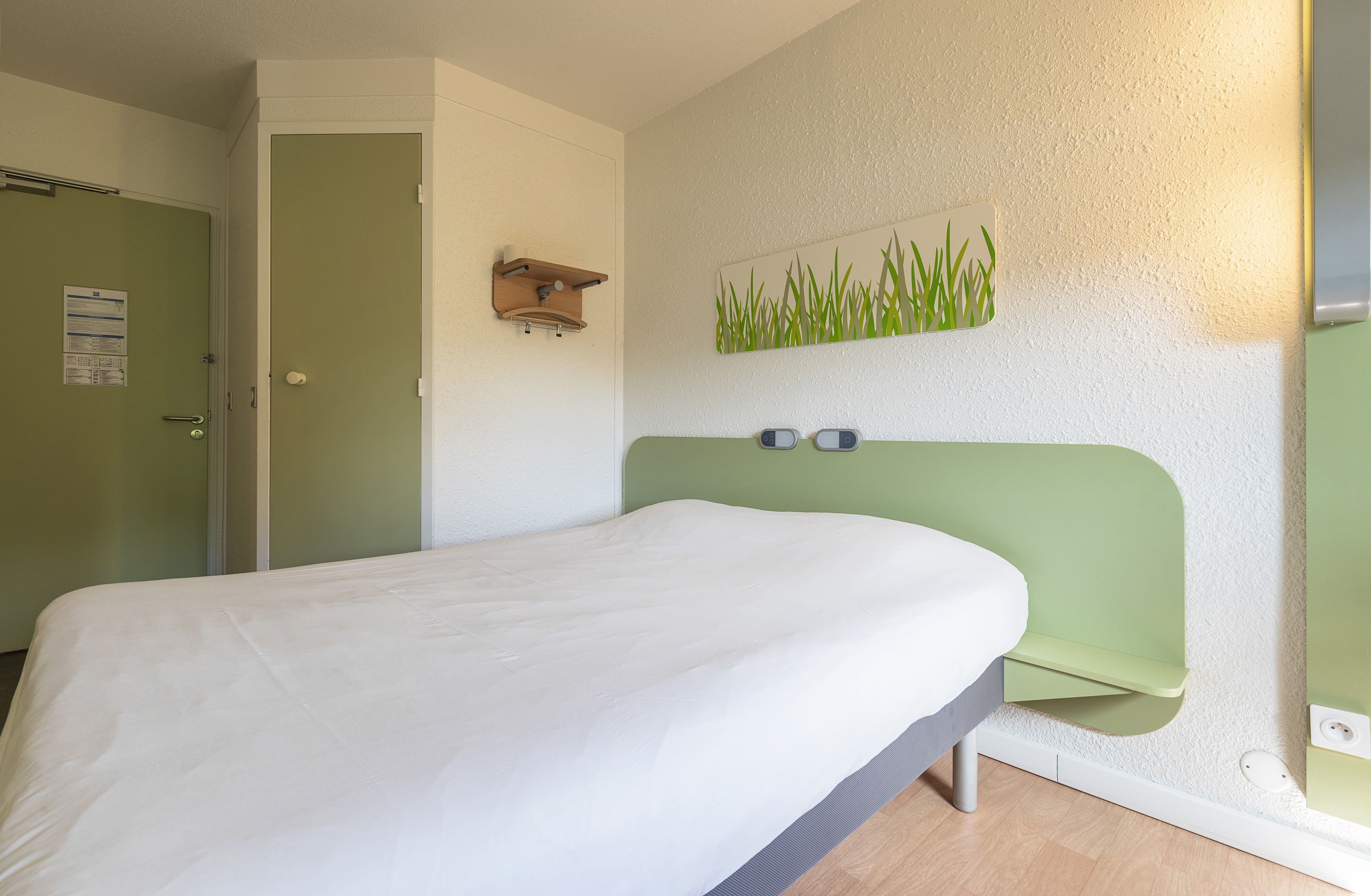 A room with a bed, a green headboard, a wall-mounted shelf, and a wall art piece.