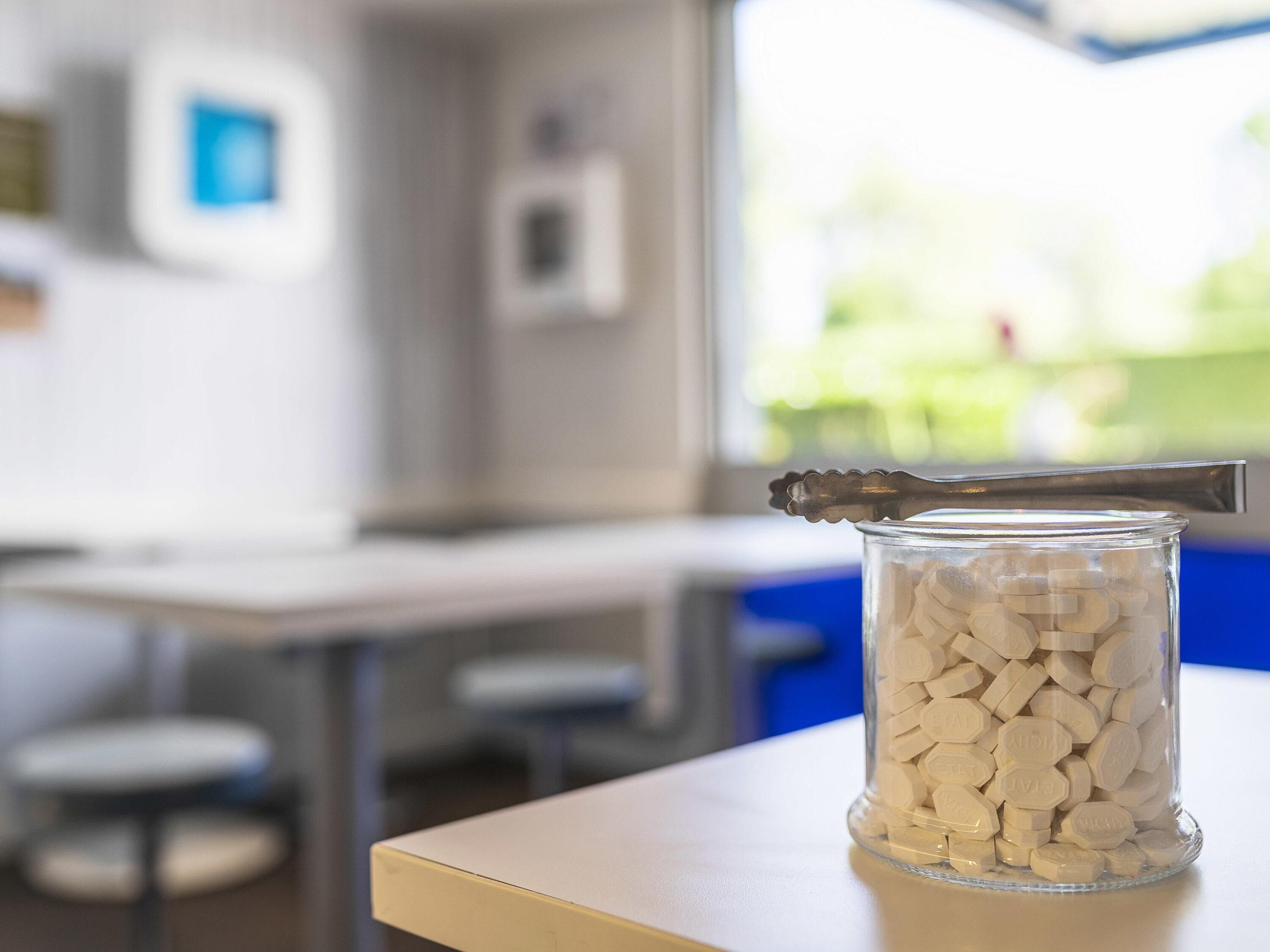 A clear glass jar filled with white pills on a table with a metal spoon resting on top.