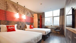 Une chambre d’hôtel avec deux lits, une petite table, une chaise et une grande fenêtre donnant sur la ville.
