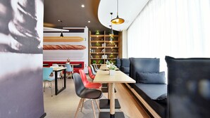 Un espace de repas moderne avec une longue table, plusieurs chaises et un canapé. On aperçoit, à l’arrière-plan, une bibliothèque agrémentée de plantes et de livres.
