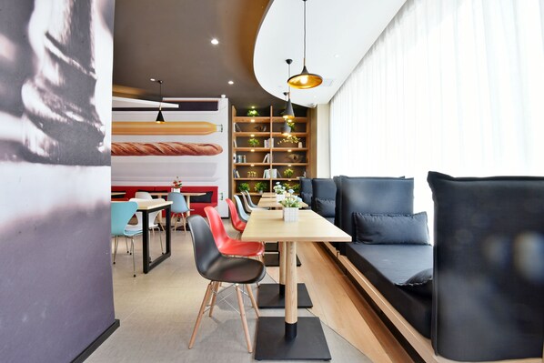 A modern dining area with a long table, multiple chairs, and a sofa. There is a bookshelf with plants and books in the background.