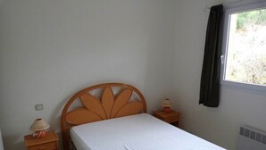 A single bed with a wooden headboard, a nightstand with a lamp, and a window with a curtain. - Mini-villa No. 4 - 2/4 people (PORTO-VECCHIO)