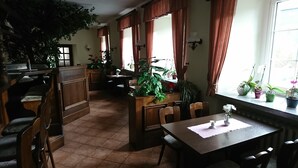 A dining area with wooden furniture, potted plants, and large windows with curtains. - Gaststätte und Pension Erbgericht (Großhartmannsdorf)
