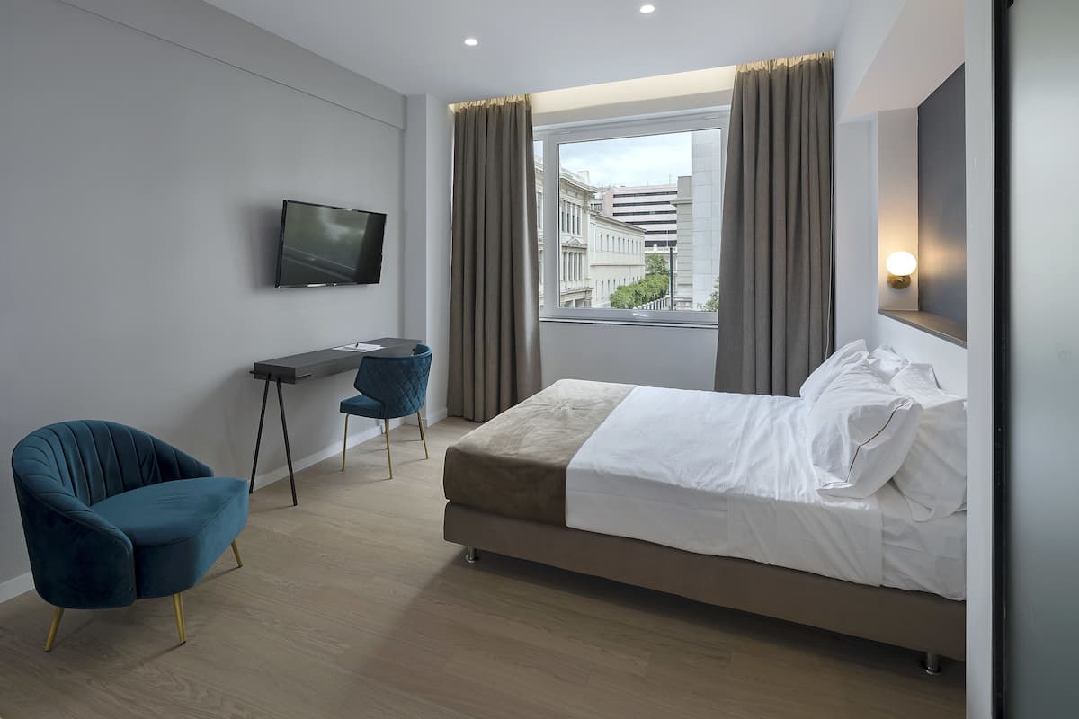 A modern hotel room with a bed, a desk, a chair, a TV, and a window with curtains.
