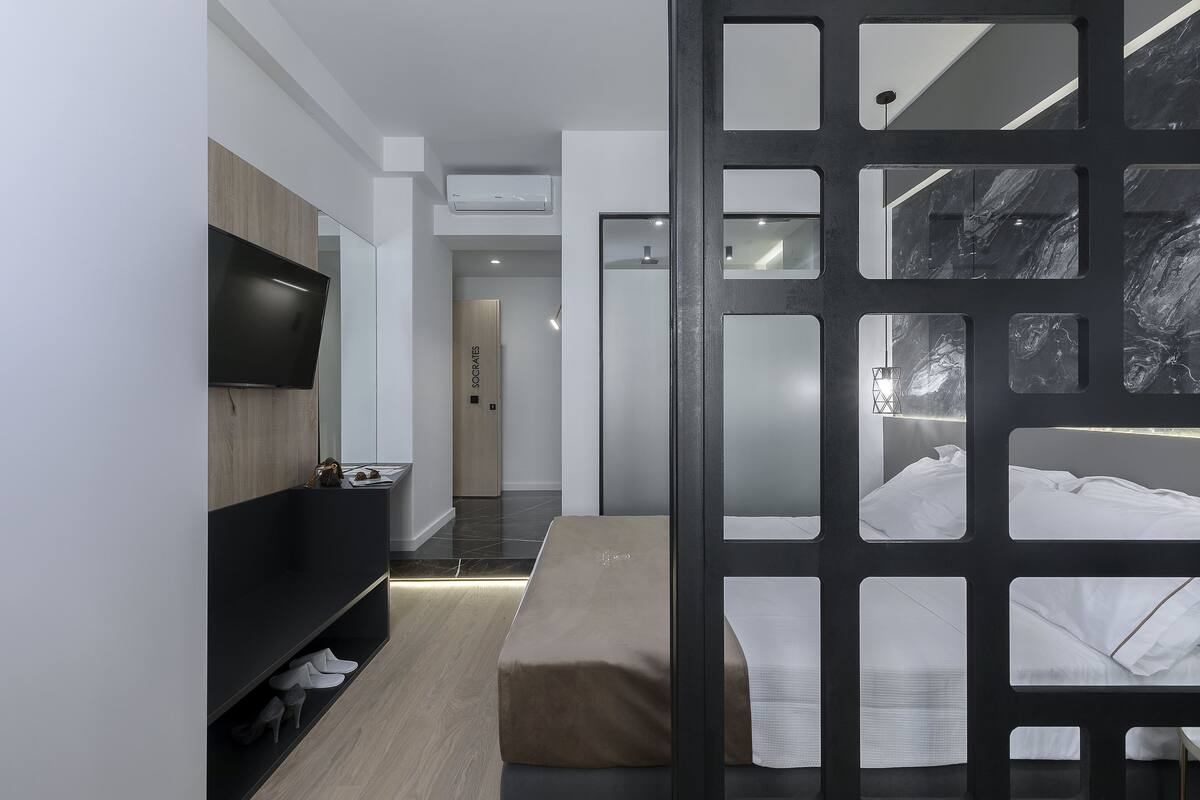 A modern hotel room with a flat-screen TV, a wooden vanity, a bench, and a bed with white linens.