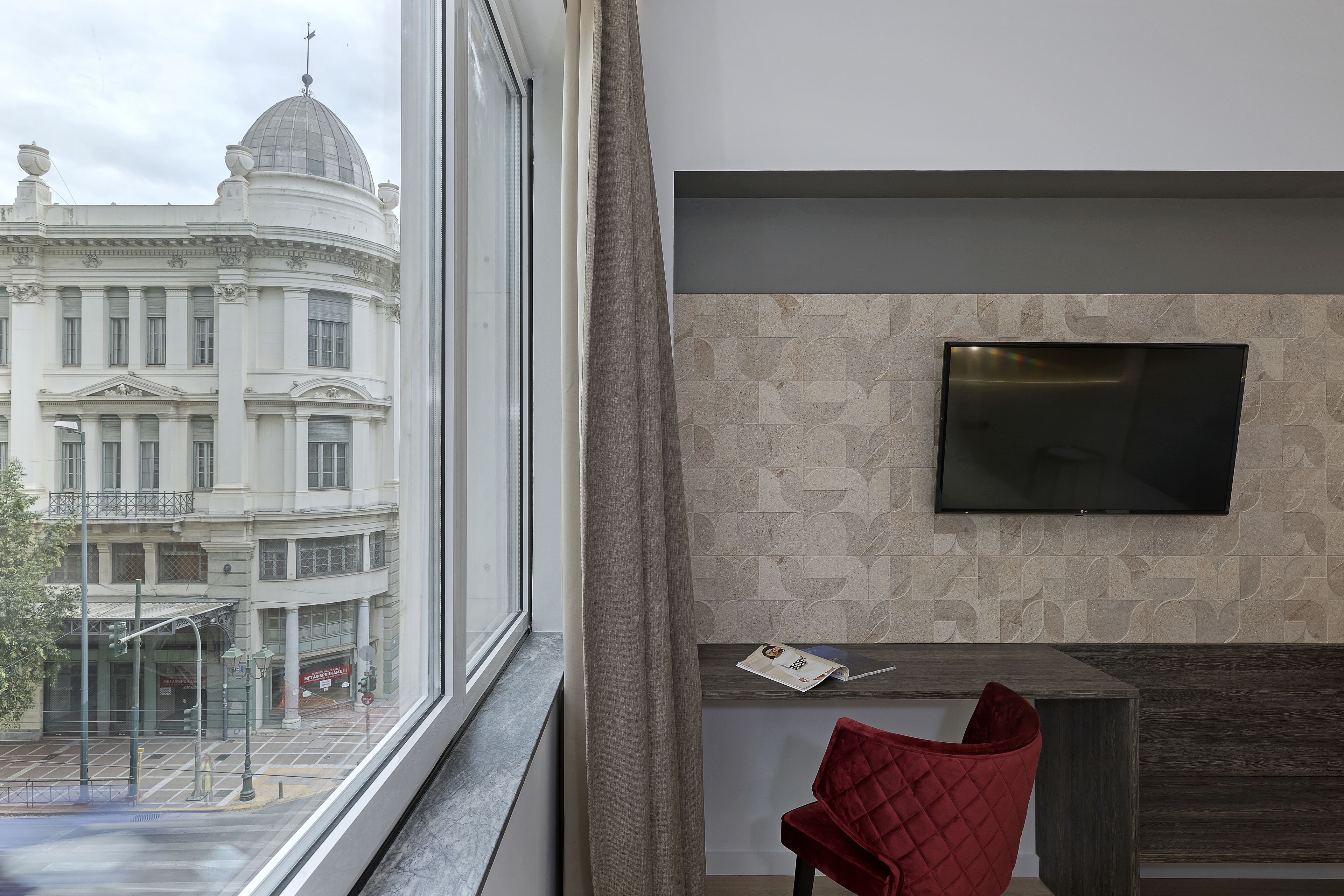 A hotel room with a large window, a flat-screen TV mounted on the wall, a red chair, and a small table.