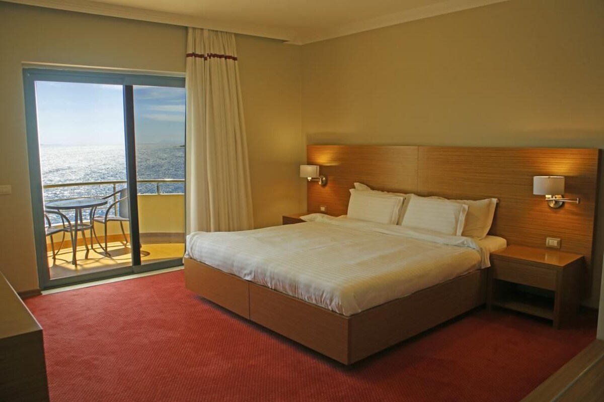 Una habitación de hotel con una cama grande, dos lámparas de noche, vistas al mar a través de una puerta corrediza y una pequeña mesa con sillas.