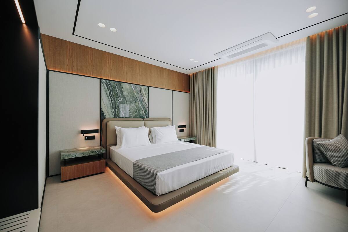 A modern hotel room with a large bed, bedside tables, a built-in bench, and a large window with curtains.