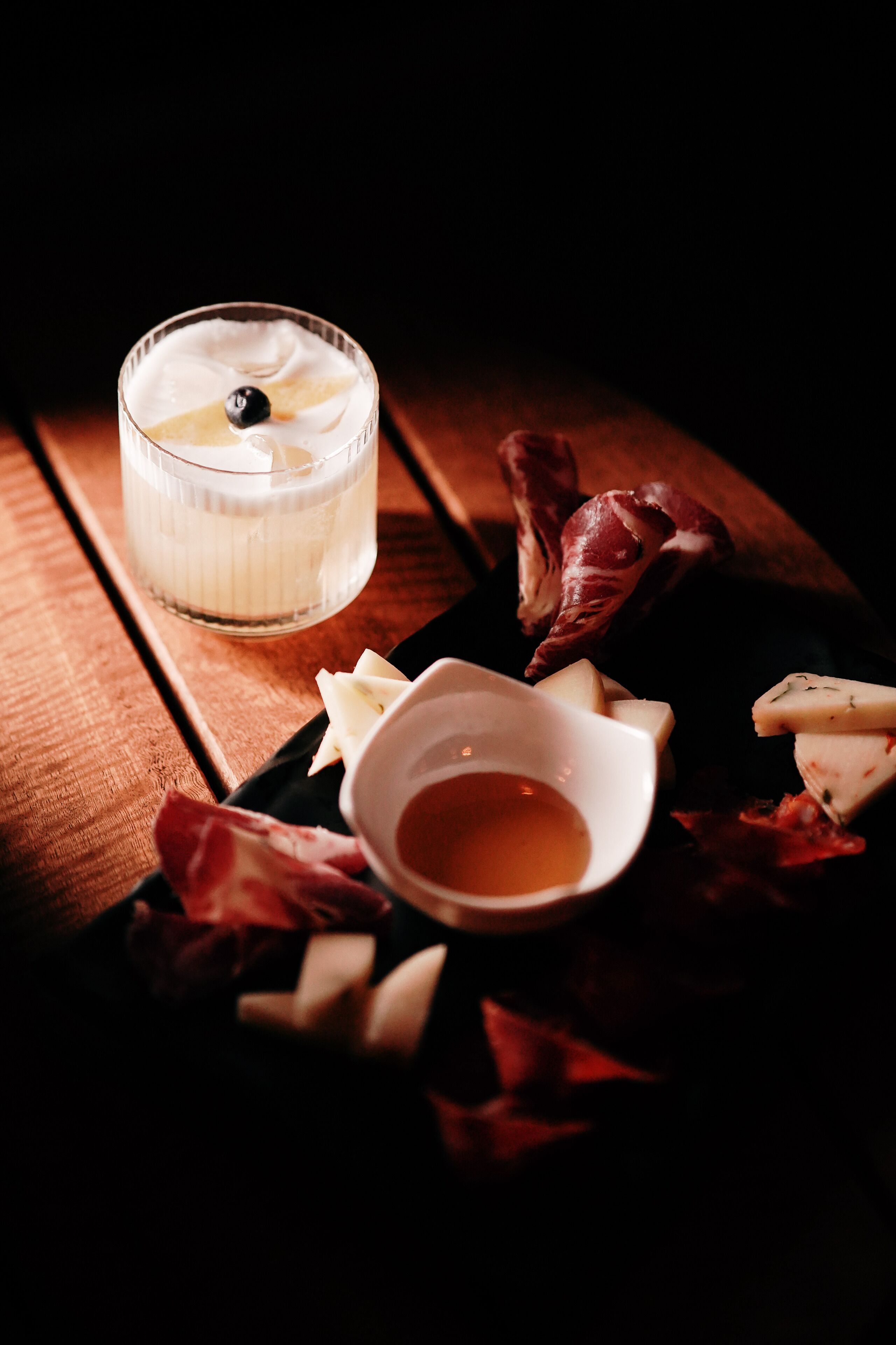 A glass of white beverage with a blueberry garnish, a small bowl of honey, and various cheeses on a wooden board.