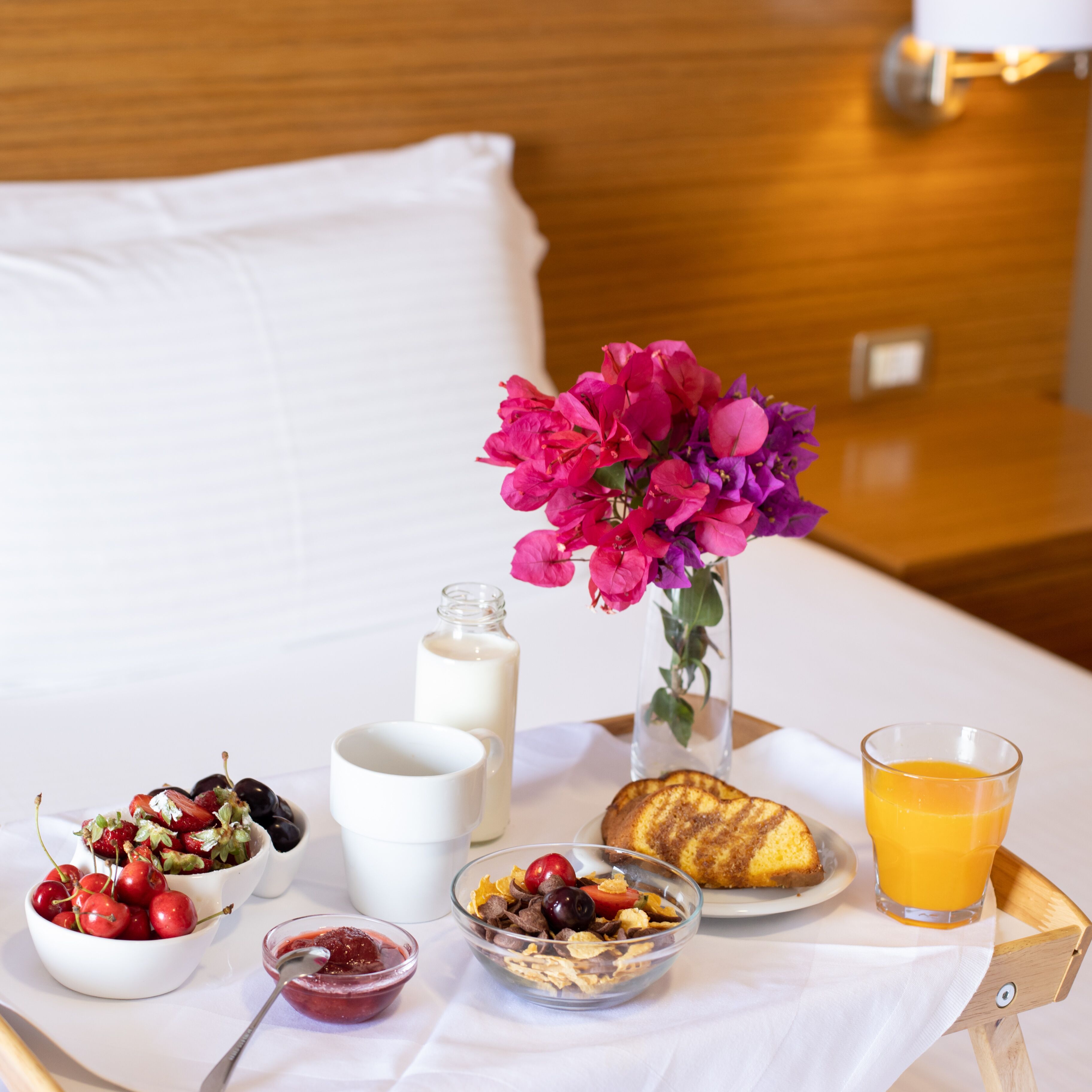 Ein ordentlich arrangiertes Frühstücksbrett mit verschiedenen Speisen, einem Glas Orangensaft und einer Blumenvase auf einem Bett.