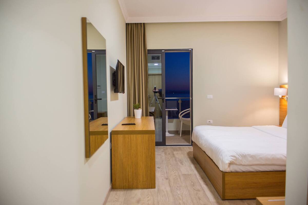 A hotel room with a bed, a desk, a chair, a TV, and a view of the outside.