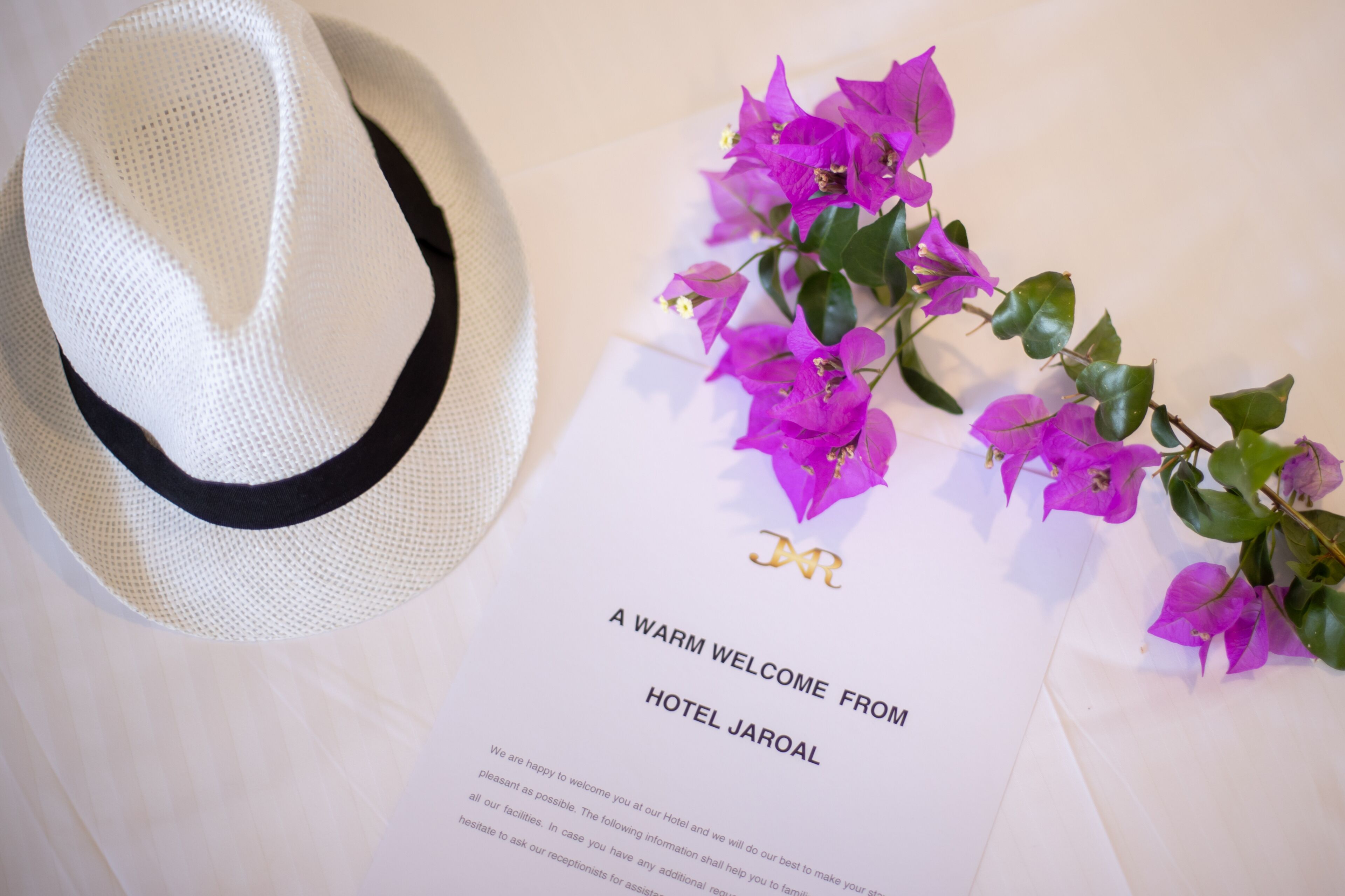Un sombrero blanco, una tarjeta de bienvenida del Hotel Jarol y flores moradas.