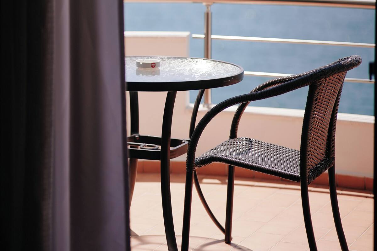 A balcony with a table and two chairs, overlooking a body of water and distant mountains.