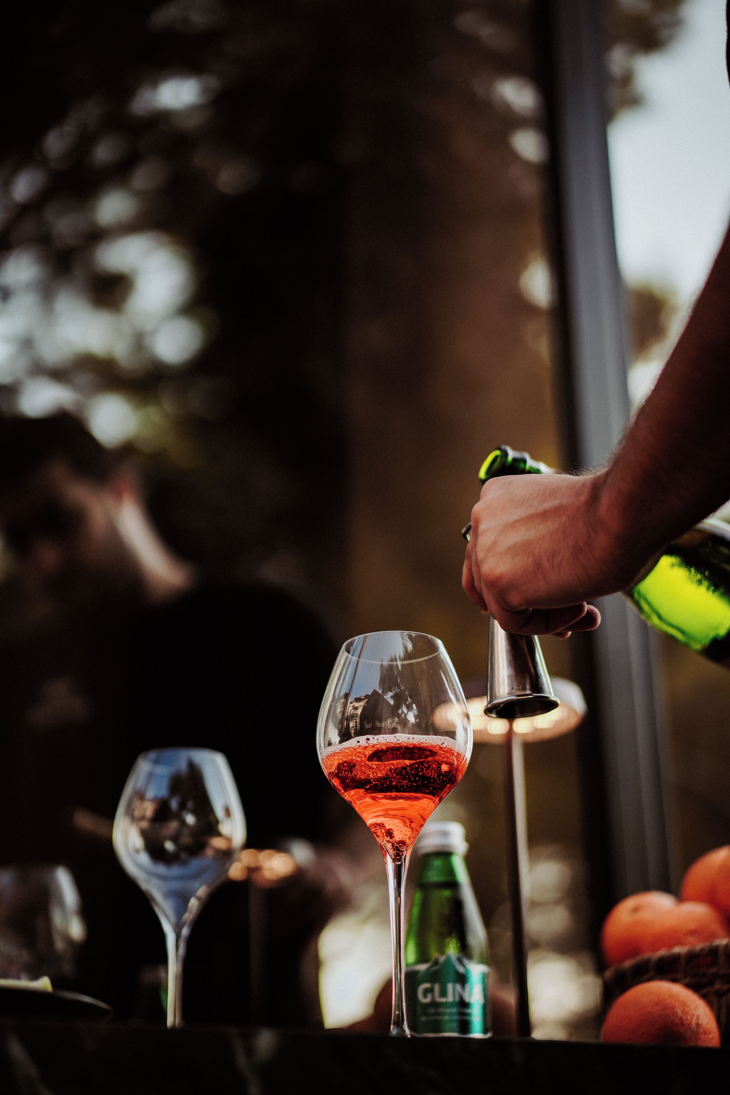 A bar setting with a person pouring a drink into a glass, a bottle of Glinat water, and oranges in the background.