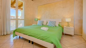 A bedroom with a green bedspread, two pillows, a white bedside table, and a nightstand with a lamp. - Fedra (Manerba del Garda)