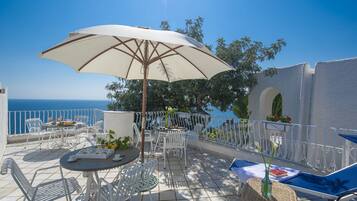 Een terras met witte metalen stoelen en tafels, een grote parasol en uitzicht op zee.