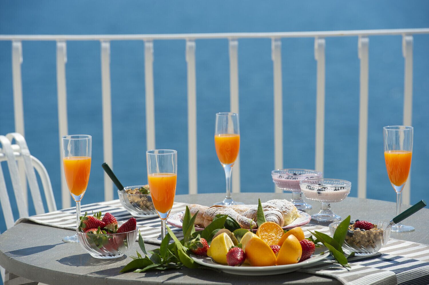 Een tafel met diverse ontbijtartikelen, zoals fruit, een kom cornflakes en glazen jus d'orange, voor een blauwe reling.