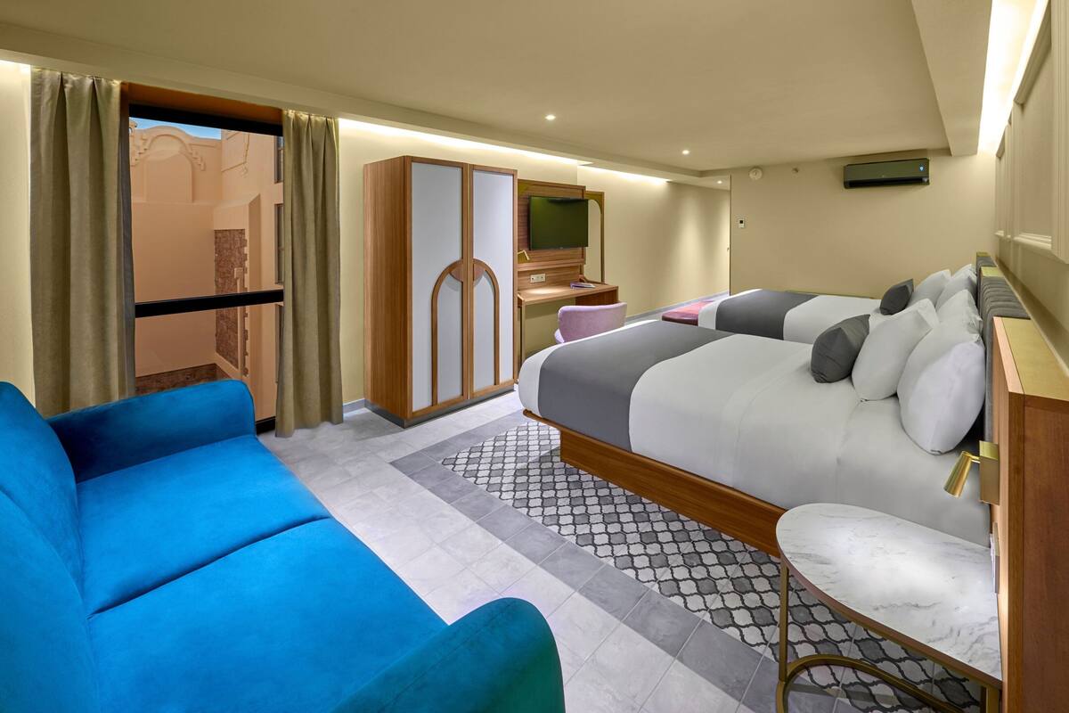 A hotel room with a large bed, a blue sofa, a wooden wardrobe, and a desk with a TV.