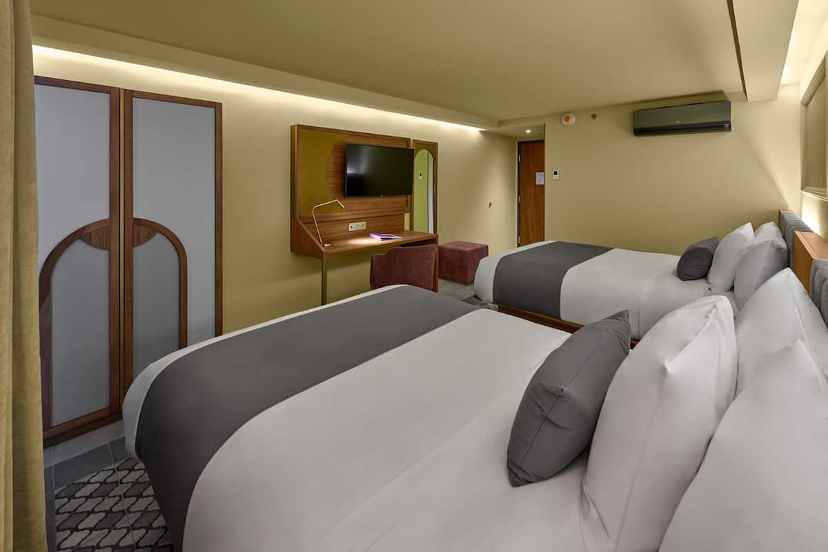 A hotel room with two beds, a desk with a chair, a television, and an air conditioning unit.