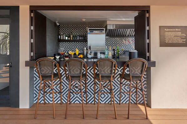 A bar area with three wicker chairs, a counter with bar tools, and a patterned backsplash.