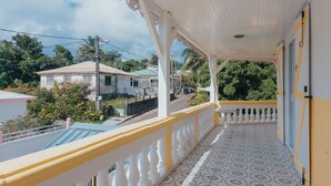Un balcón techado con suelo estampado, barandillas blancas y vistas a casas y vegetación circundantes.